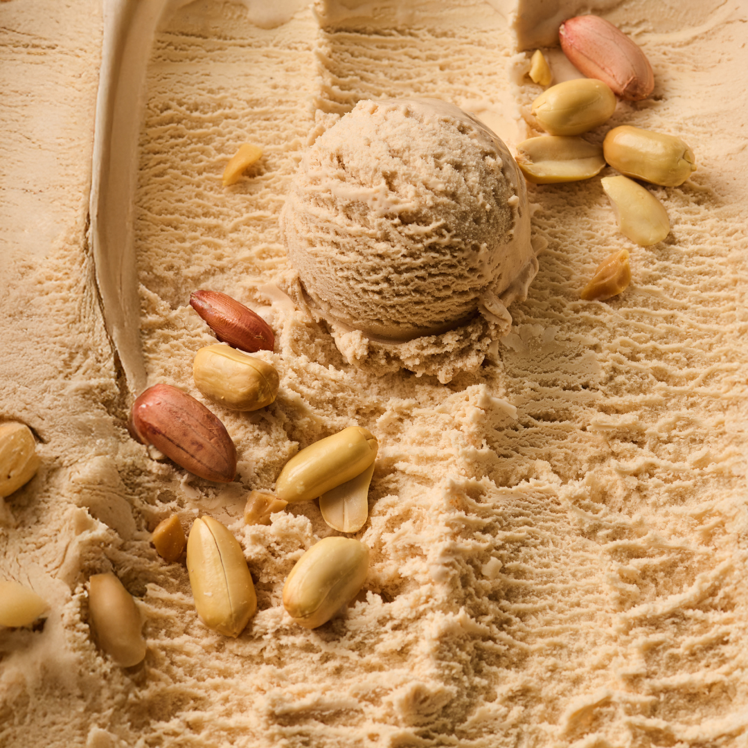 Eine Kugel Erdnusseis liegt auf einer Oberfläche aus cremigem Eis, umgeben von ganzen Erdnüssen.