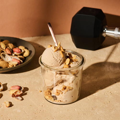 Ein Glas mit Erdnusseis und Erdnüssen, daneben ein Teller mit Schalenfrüchten auf einem Tisch mit Hantel.