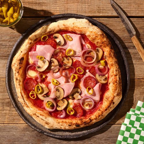 Pizza mit Schinken, Salami, Champignons, Zwiebeln und Peperoni auf einem Holzbrett, daneben ein Glas mit Peperoni.