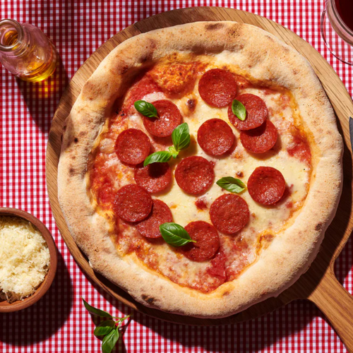 Pizza mit Salami und Basilikum auf Holzbrett, eingerahmt von Tischdecke, Parmesan und Olivenöl.