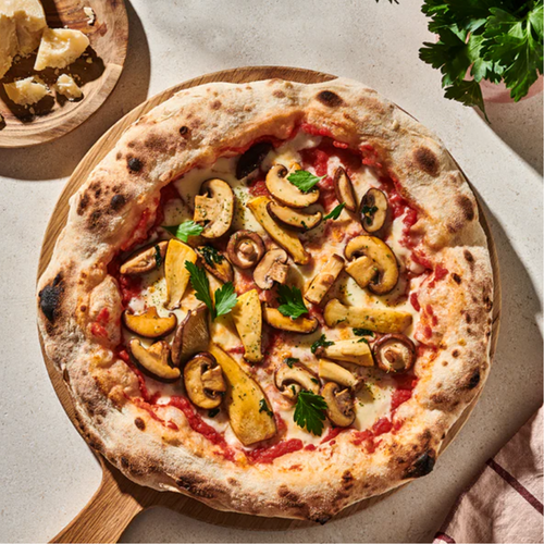 Pizza mit Pilzen und Petersilie auf einem Holzbrett, daneben Käse und frische Kräuter auf einem Tisch.