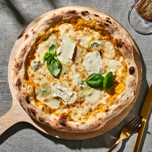Pizza mit Käse und Basilikum auf Holzbrett, neben goldenem Besteck und Weinglas auf grauem Tischtuch.