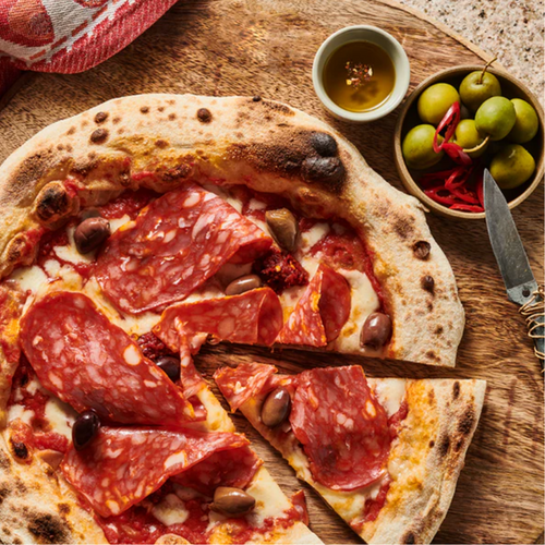 Pizza mit Salami und Oliven auf einem Holzbrett, daneben eine Schale mit Oliven und ein kleines Gefäß mit Öl.