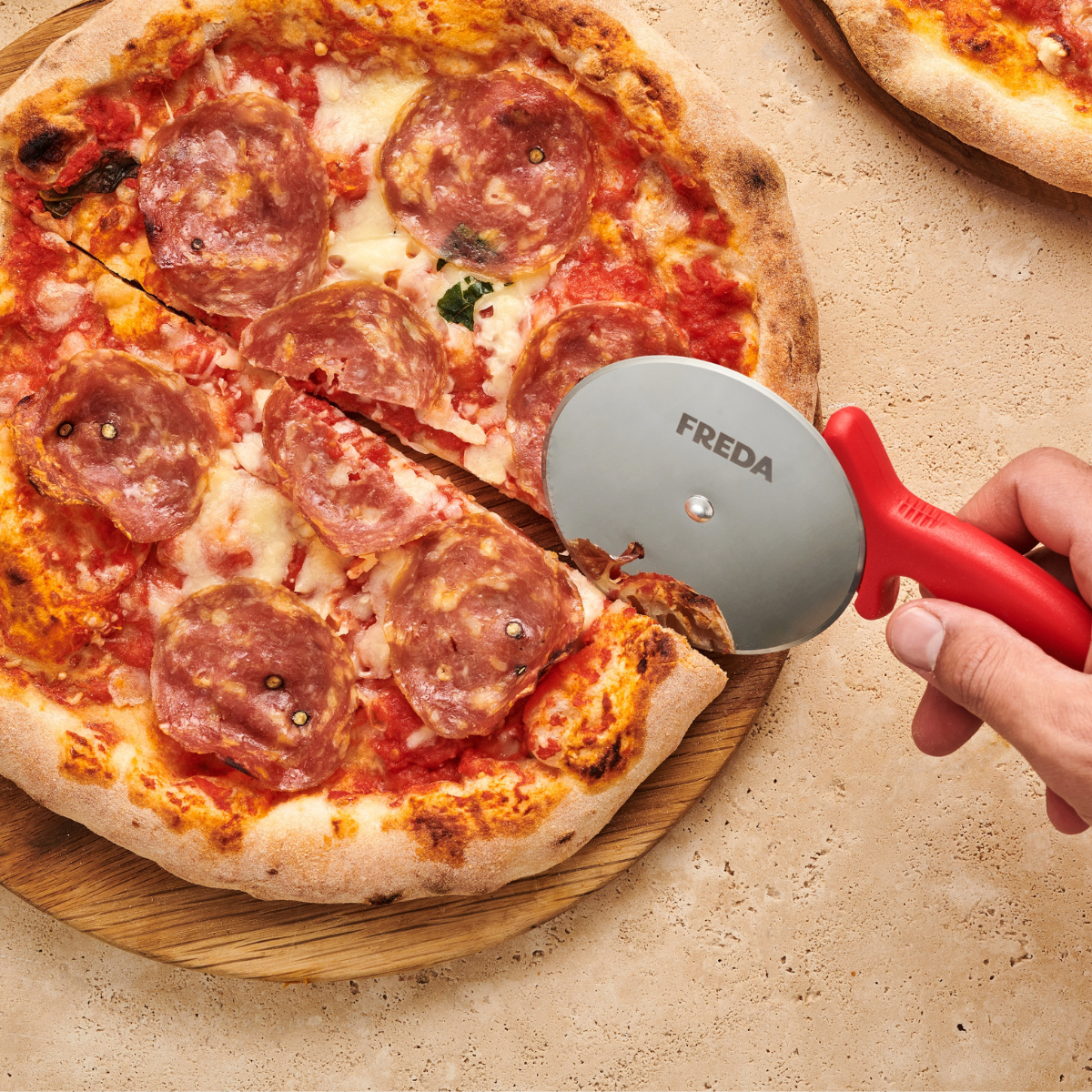 Hand schneidet eine Salami-Pizza mit einem Pizzaschneider mit rotem Griff auf einem hölzernen Schneidebrett.