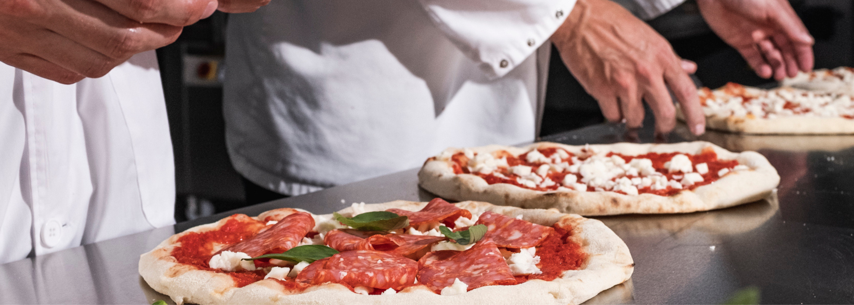 Köche belegen Pizzen mit Tomatensoße, Käse, Salami und Basilikum auf einer Edelstahloberfläche.