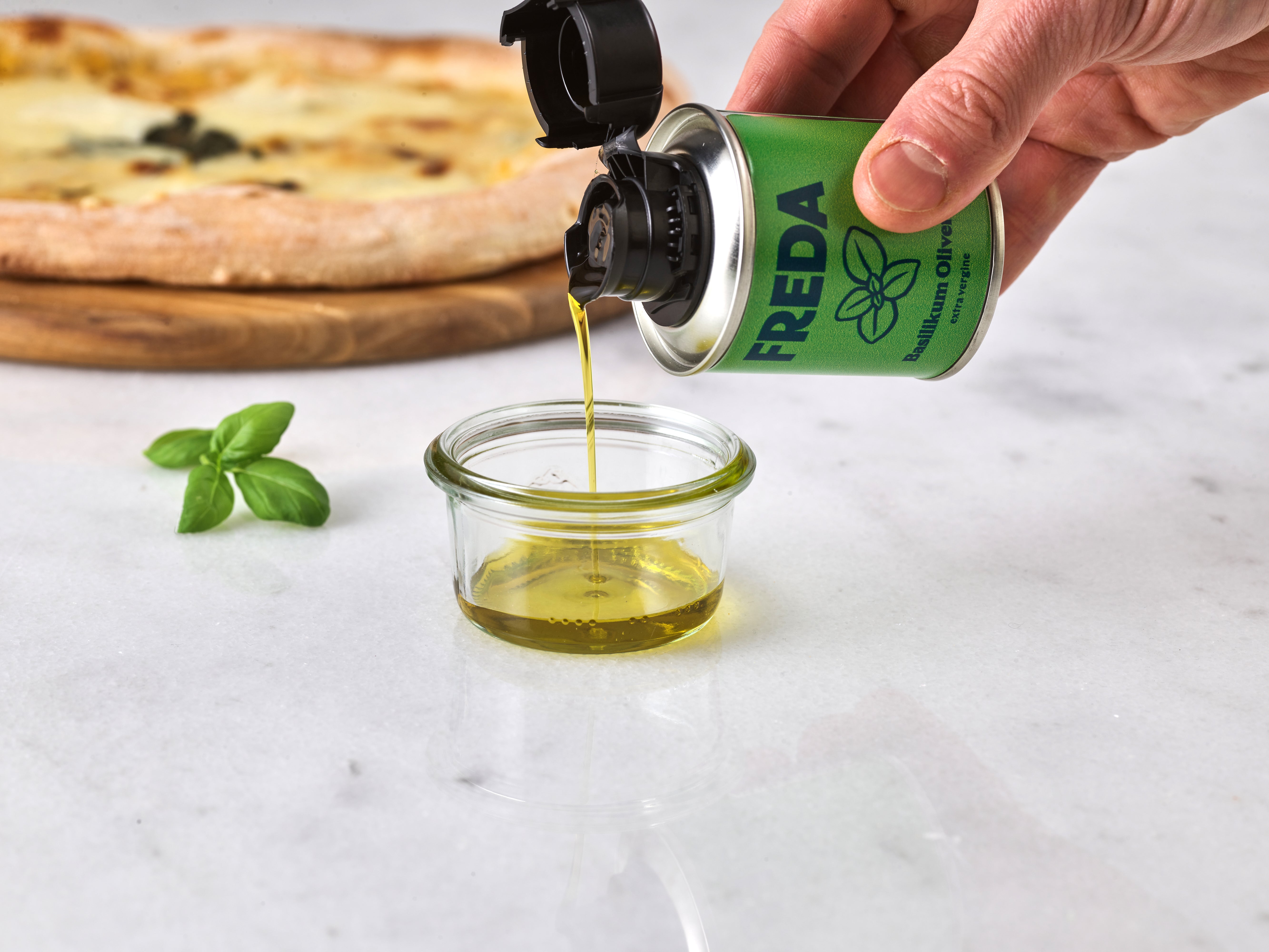 Eine Hand gießt Öl aus einer grünen Dose in ein Glas, daneben liegt Basilikum, im Hintergrund eine Pizza.