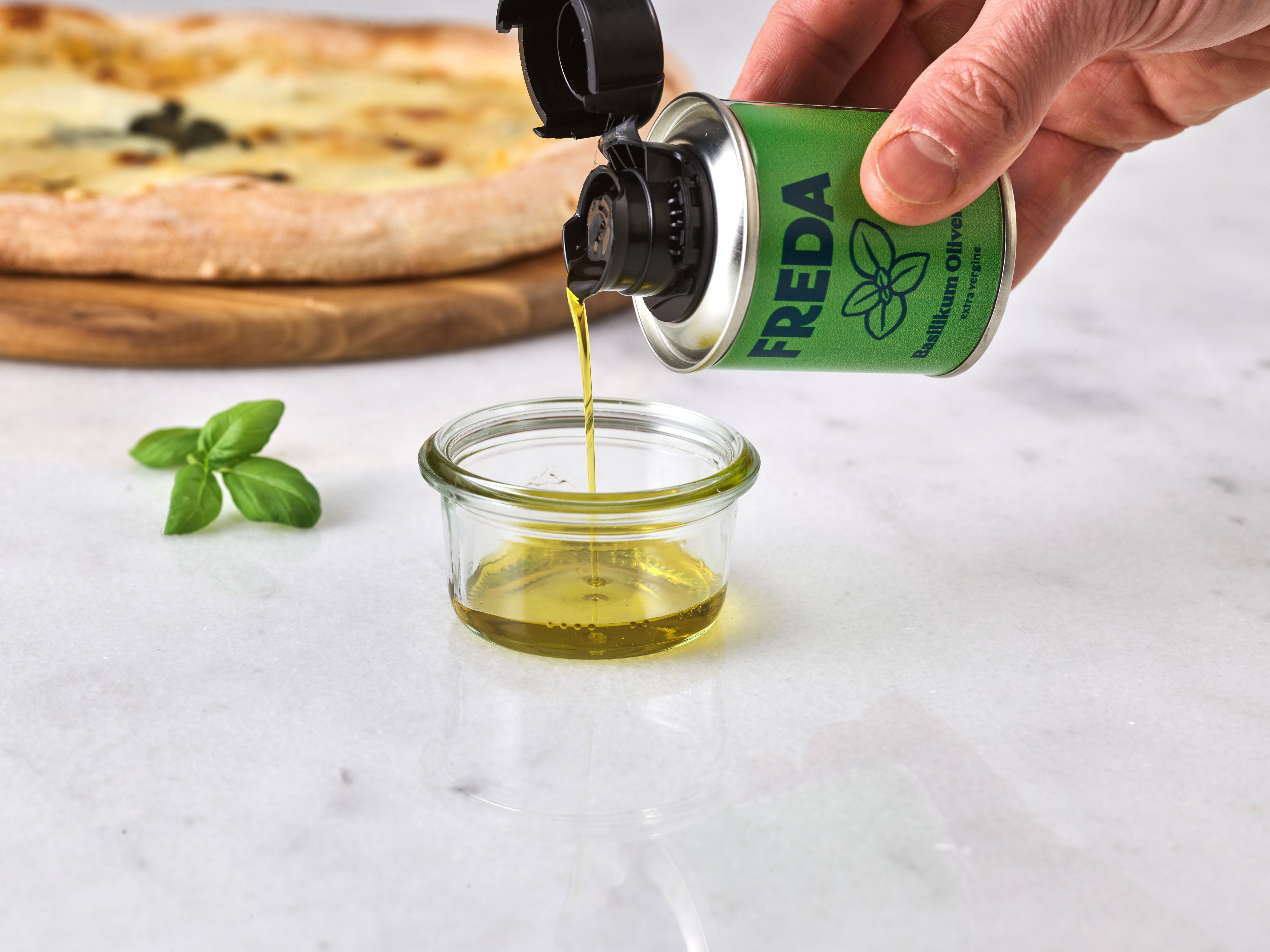 Eine Hand gießt Öl aus einer grünen Dose in ein Glas, daneben liegt Basilikum, im Hintergrund eine Pizza.