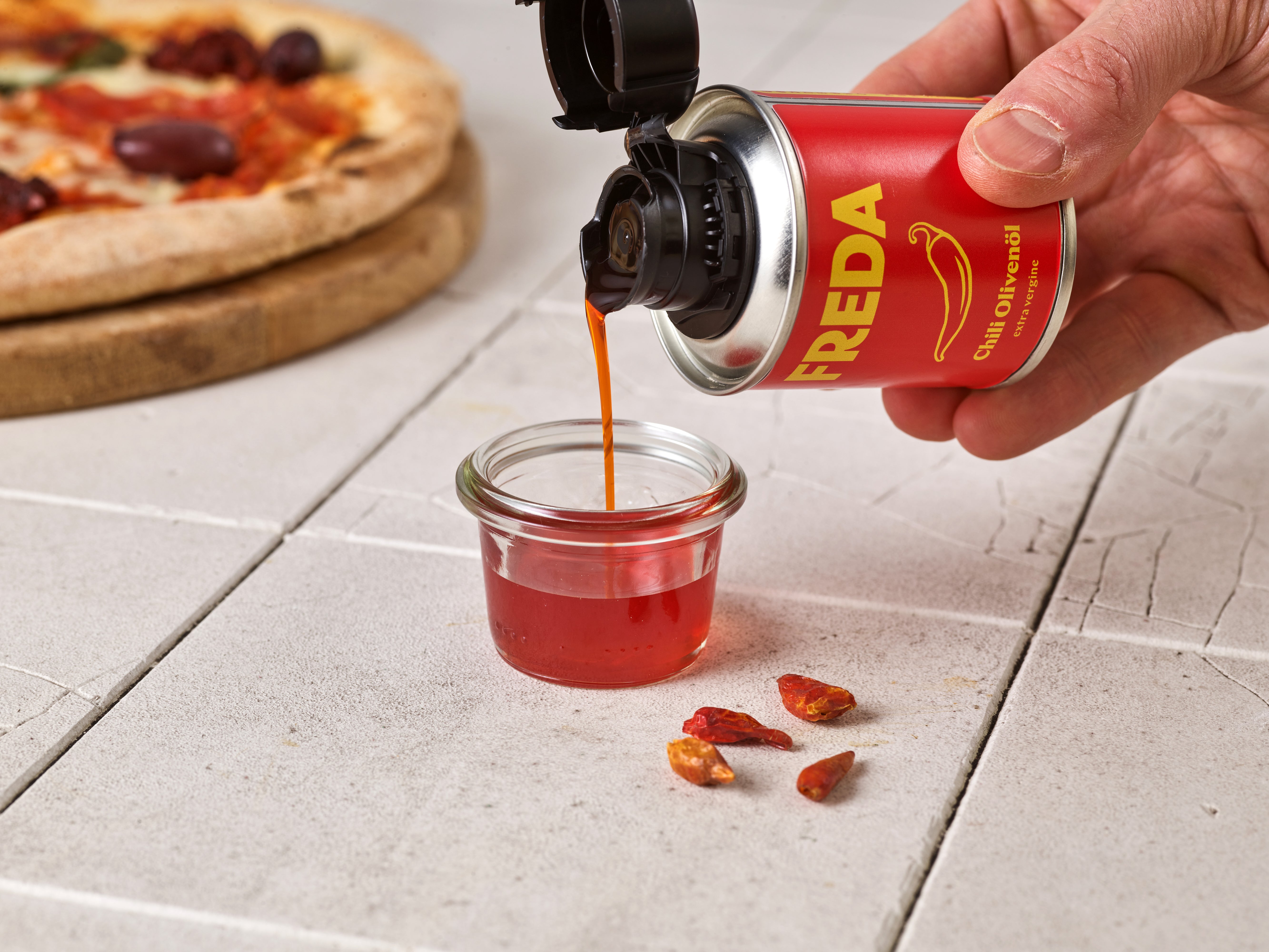 Hand gießt Chili-Olivenöl aus einer Dose in ein Glas, mit Pizza und Chilischoten im Hintergrund.