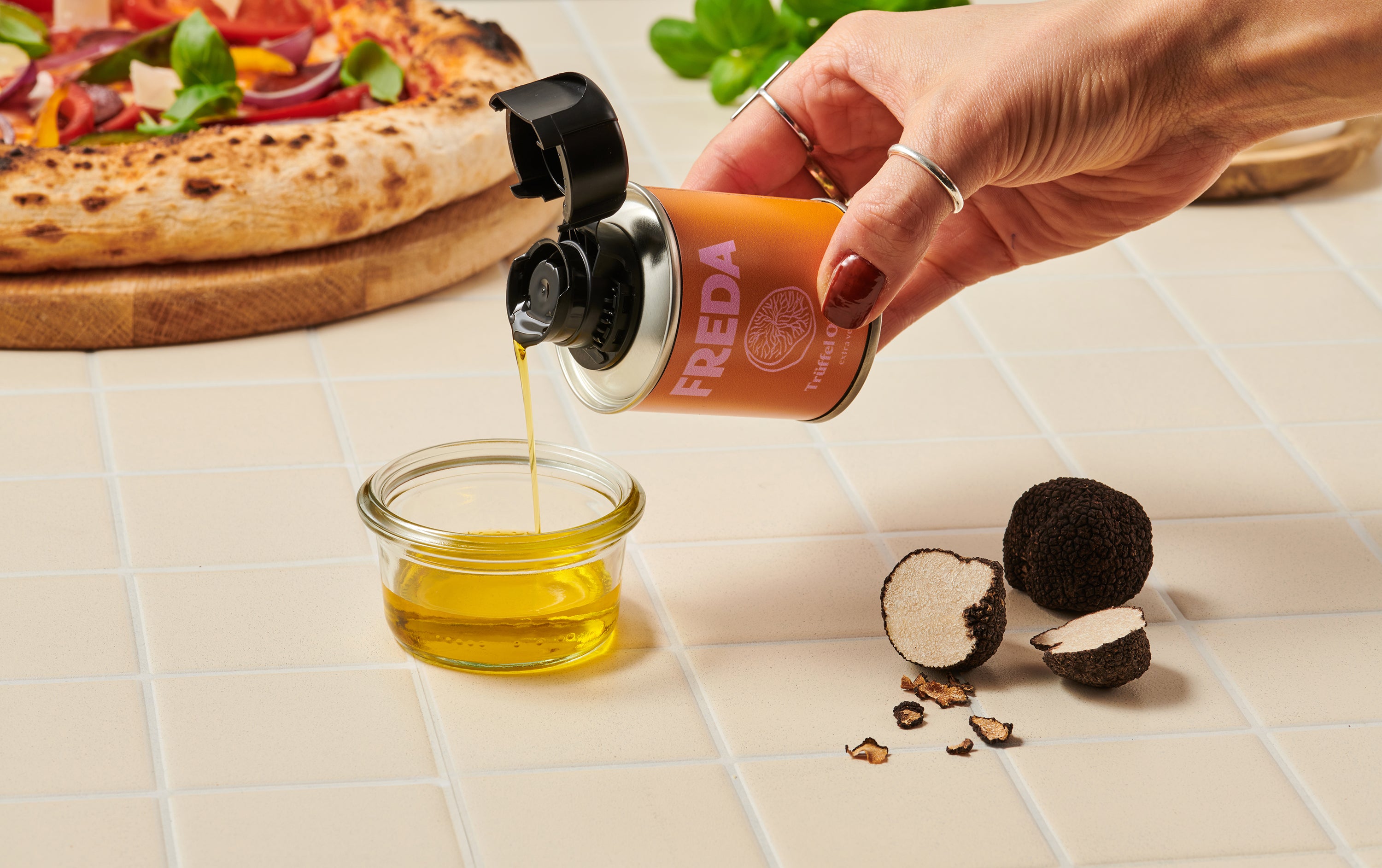 Eine Hand gießt Trüffelöl aus einer kleinen Dose in ein Glasgefäß neben einer Pizza und frischen Trüffeln.