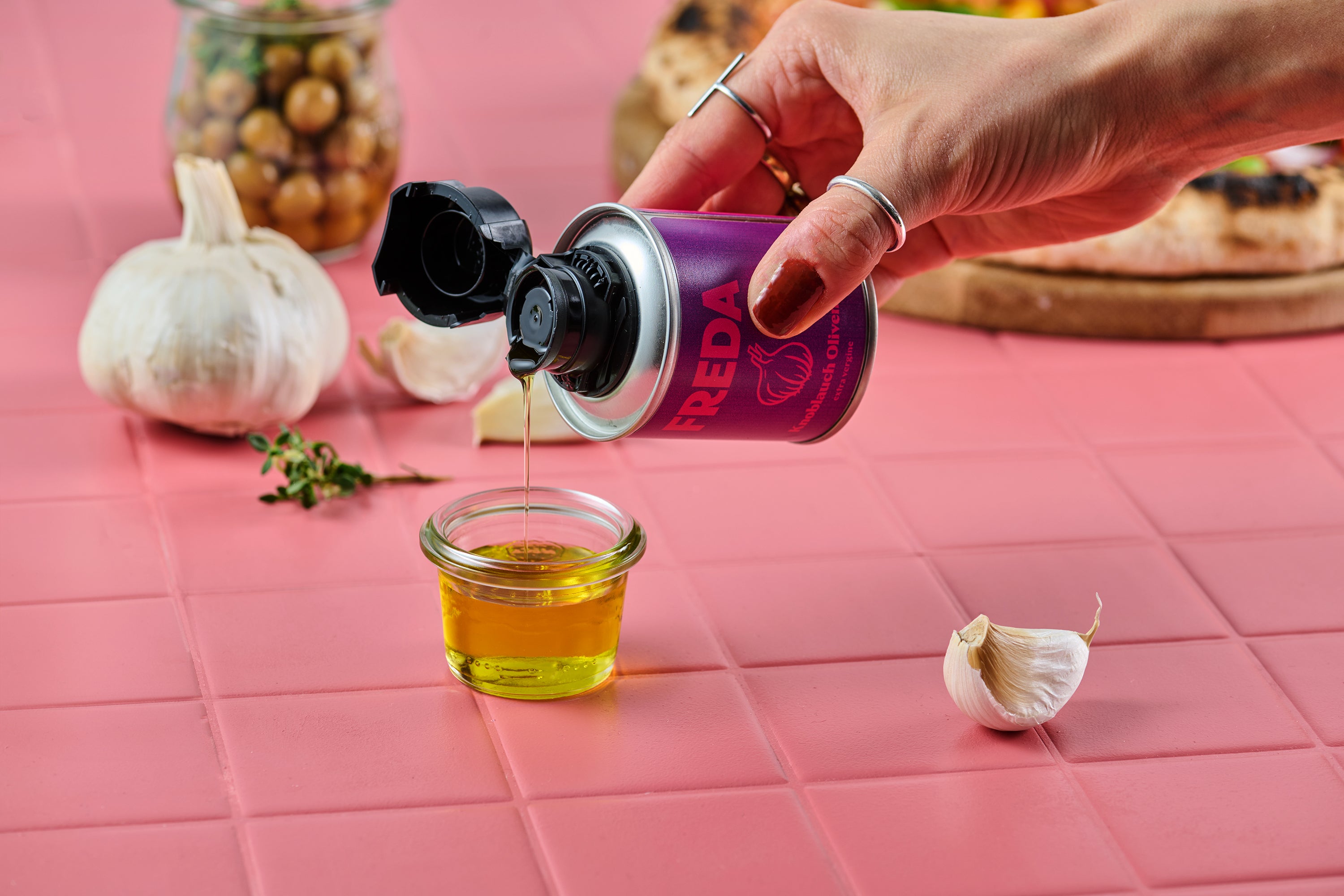 Hand hält eine Dose und gießt Öl in ein Glas, umgeben von Knoblauch und Kräutern auf einem rosa Tisch.