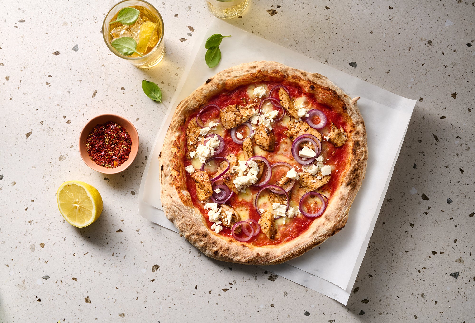 Pizza mit Hähnchen, roten Zwiebeln und Feta auf einem weißen Untergrund, daneben Pfefferflocken, ein Zitronenstück und getränke.