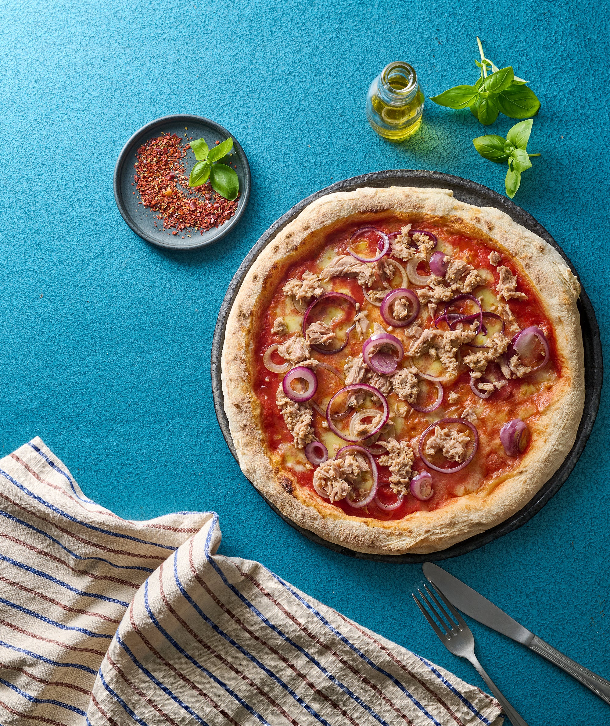 Pizza mit Thunfisch und roten Zwiebeln auf einem blauen Tisch, daneben Chili, Basilikum, Öl und ein gestreiftes Tuch.