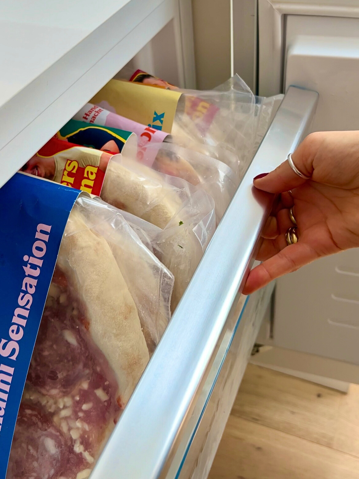 Eine Hand öffnet eine Gefrierschublade mit verpackten Tiefkühllebensmitteln, darunter eine Pizza Salami.