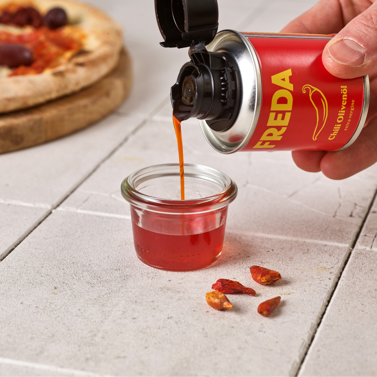 Eine Hand gießt Chili-Öl aus einer roten Dose in ein Glasgefäß, im Hintergrund liegt eine Pizza.