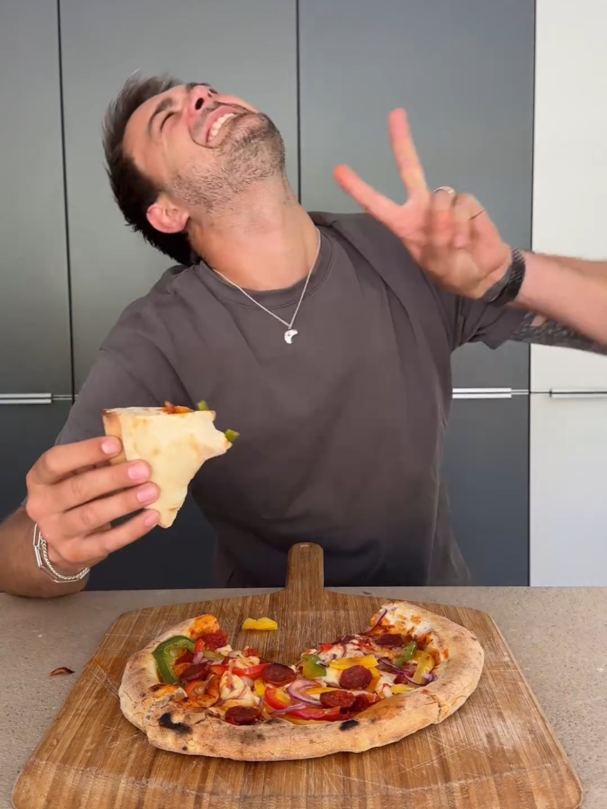 Ein Mann lächelt und zeigt ein Peace-Zeichen, während er ein Pizzastück in der Hand hält; vor ihm liegt eine Pizza.