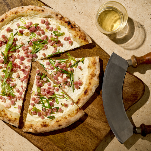 Pizza mit Sahnesoße, Schinken und Frühlingszwiebeln auf Holzbrett, mit Halbmond-Messer und Glas Weißwein daneben.