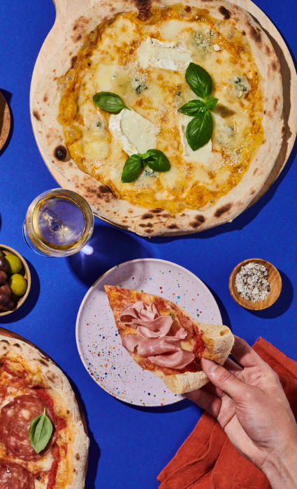 Hand hält Pizzastück über Teller, umgeben von Pizzen, Oliven, Gewürzen und einem Glas Wein auf blauem Tisch.