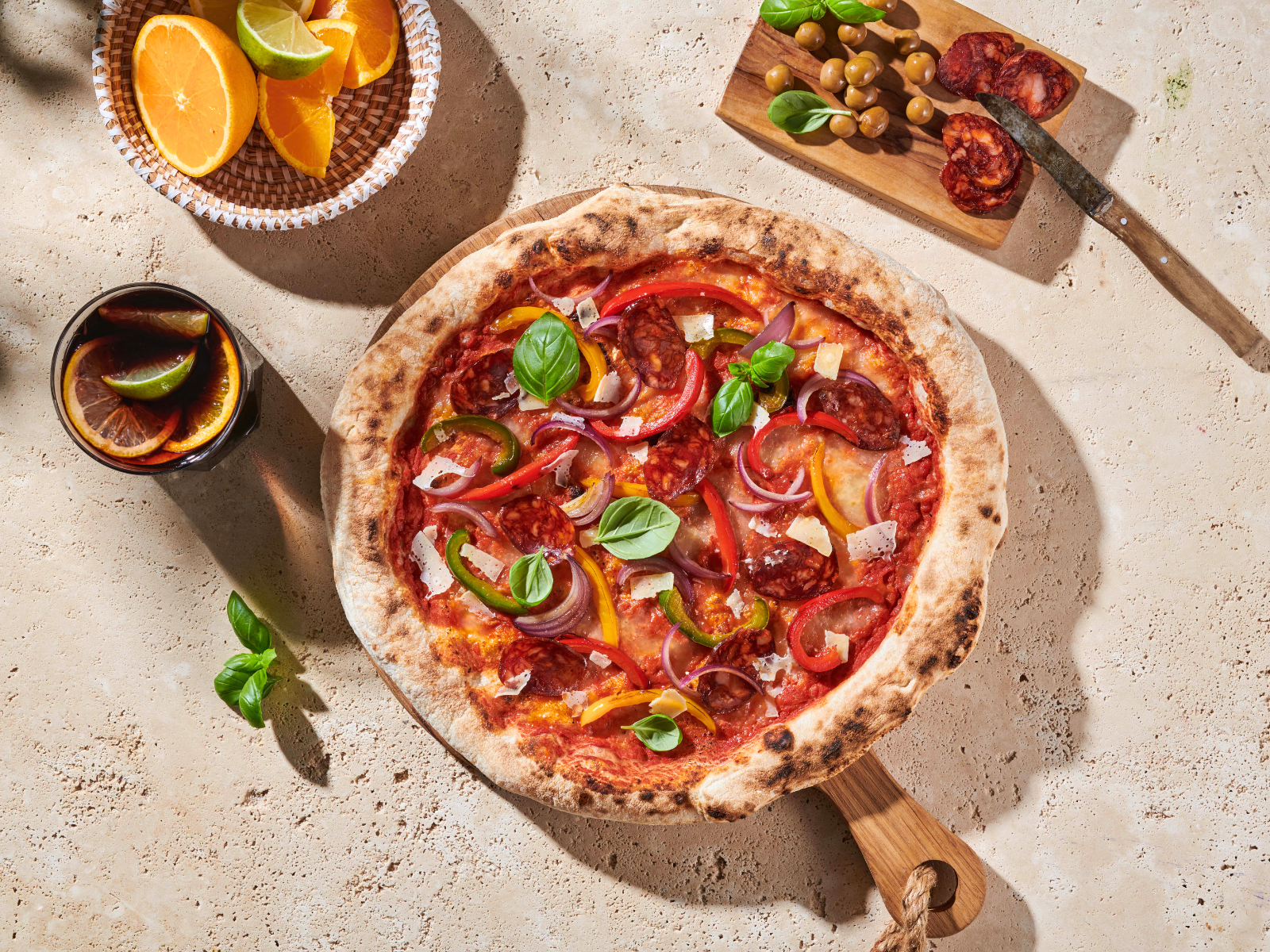 Pizza mit Paprika und Basilikum auf einem Holzbrett, daneben ein Getränk mit Zitrusfrüchten und ein Holzbrett mit Oliven.