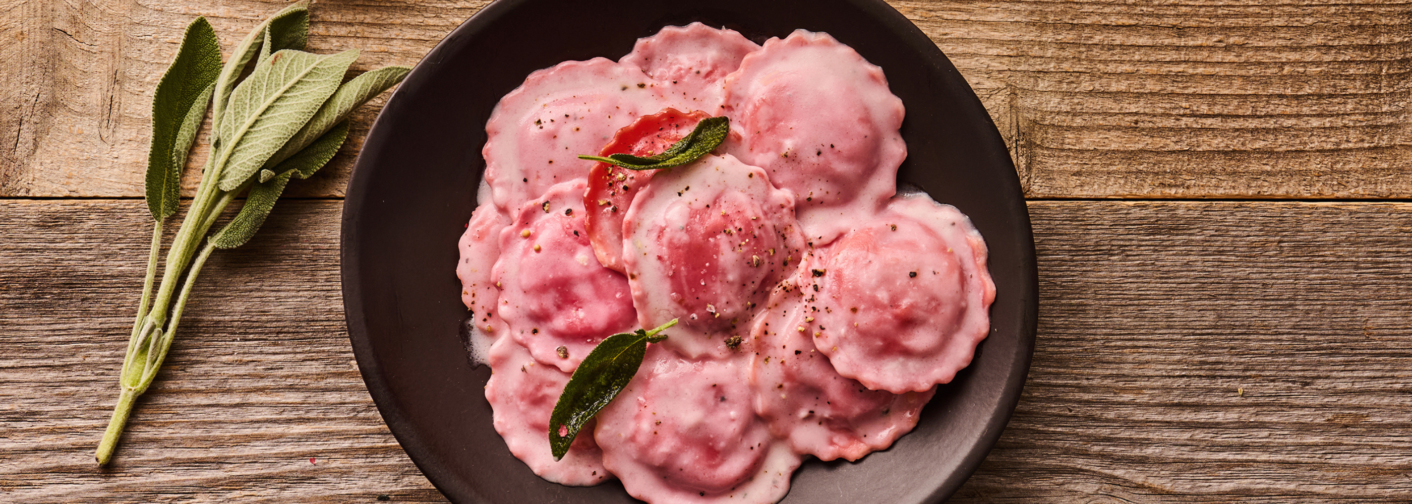 Rosa Ravioli mit Sahnesauce und Salbeiblättern auf einem schwarzen Teller, daneben frischer Salbei auf Holztisch.