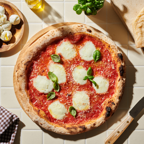 Pizza Margherita auf Holzbrett, umgeben von Mozzarella, Olivenöl, Basilikum und Gebäck auf einem Tisch mit Messer.