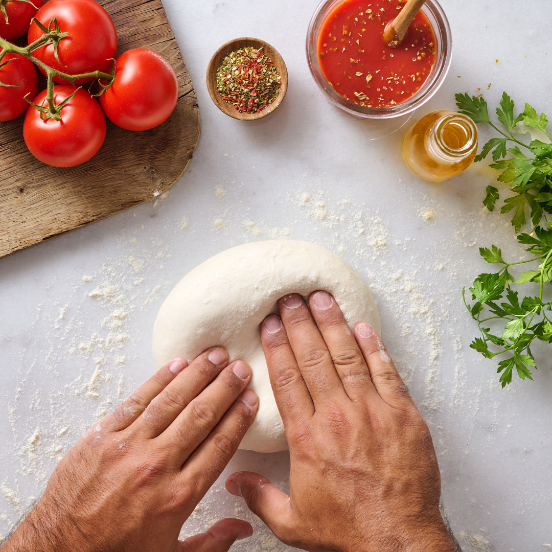 Hände kneten Pizzateig auf bemehlter Oberfläche, umgeben von Tomaten, Kräutern, Tomatensauce und Ölflasche.