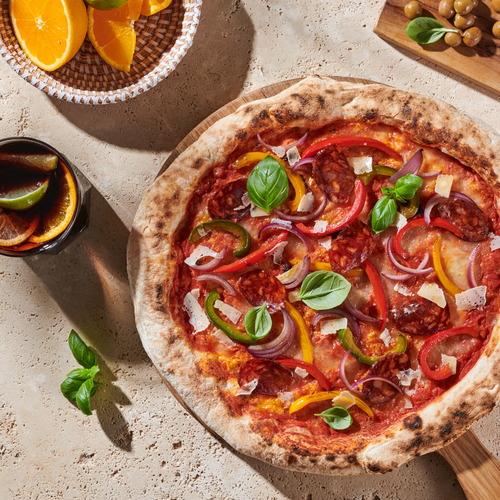 Pizza mit Peperoni, Zwiebeln, Basilikum und Käse auf einem Tisch, daneben ein Getränk mit Zitrusfrüchten und Oliven.