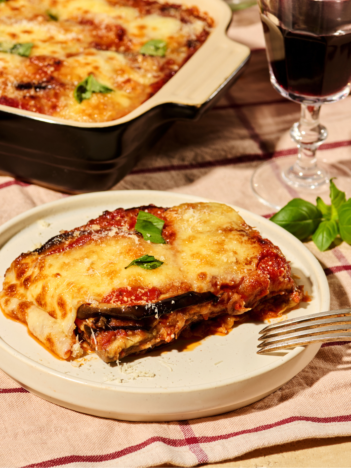 Ein Teller Auberginenauflauf mit geschmolzenem Käse und frischen Kräutern, daneben ein Glas Rotwein.
