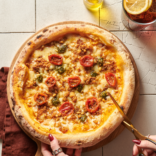 Pizza mit Tomaten, Pesto und Pinienkernen auf einem Holzbrett, eine Person schneidet ein Stück ab.