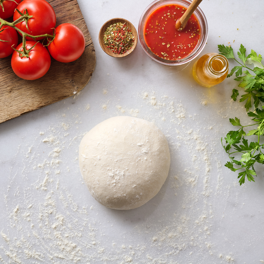 Teigkugel auf bemehlter Fläche, umgeben von Tomaten, Kräutern, Tomatensauce und Öl.