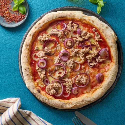Eine Pizza belegt mit Thunfisch, roten Zwiebeln und Tomatensauce liegt auf einem Tisch mit Basilikum und Gewürzen.