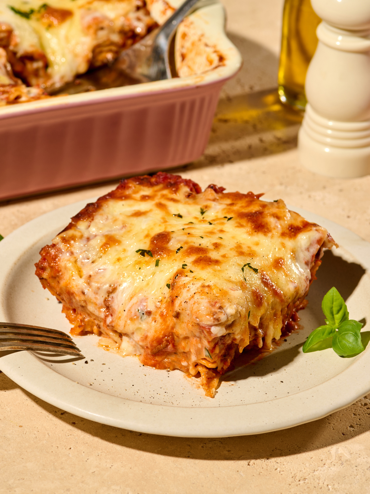 Ein Stück Lasagne mit geschmolzenem Käse auf einem Teller, daneben Basilikum. Im Hintergrund eine Auflaufform.