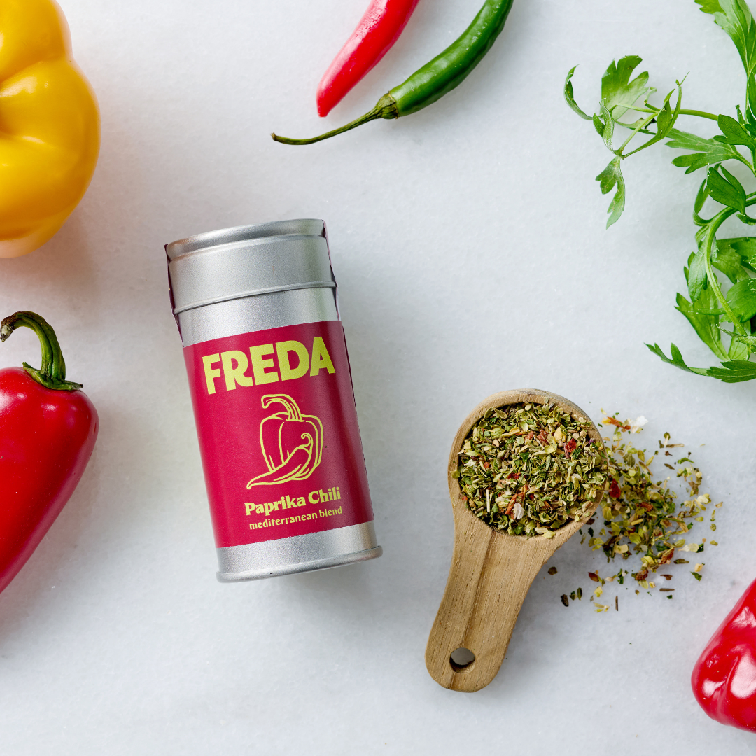 Dose mit Paprika-Chili-Gewürz, umgeben von buntem Gemüse und einem Holzlöffel mit getrockneten Kräutern.