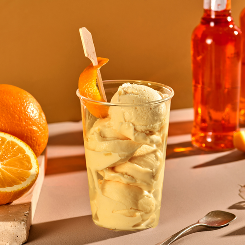 Ein Glas Vanilleeis mit einer Orangenspirale dekoriert, daneben eine Flasche Orangensirup und geschnittene Orangen.