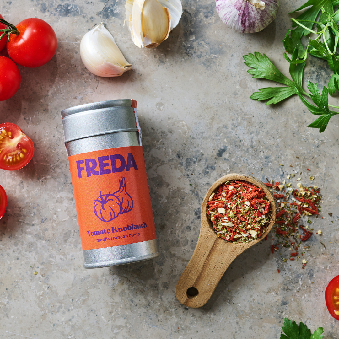 Gewürzdose "FREDA" mit Tomaten, Knoblauch, Kräutern und Gewürzen auf einer Steinoberfläche arrangiert.