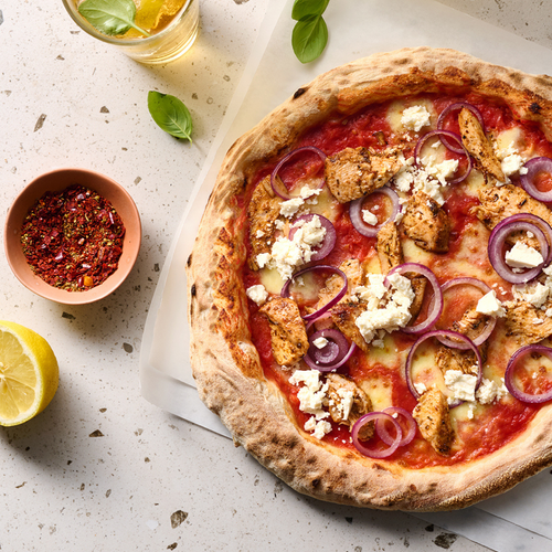 Pizza mit Hähnchen, roten Zwiebeln und Fetakäse, daneben Gewürzschale, Zitronenscheibe, Basilikumblätter und Getränk.