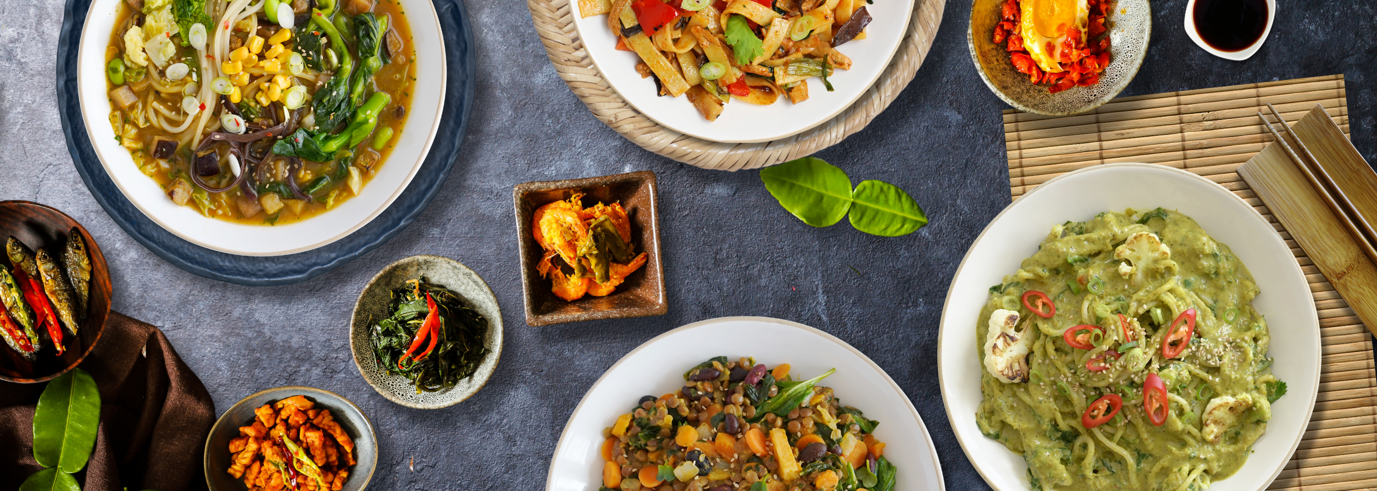 Verschiedene asiatische Gerichte in Schüsseln und Tellern, darunter Nudeln und Gemüse, auf einem Tisch arrangiert.