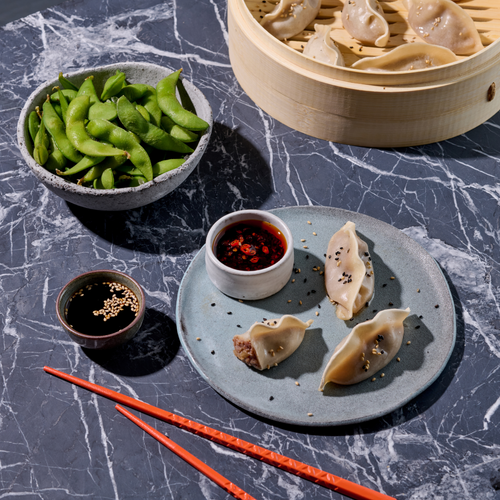 Dampfnudeln und Edamame auf Marmortisch, serviert mit Sojasauce und Chili-Dip, rote Essstäbchen daneben.