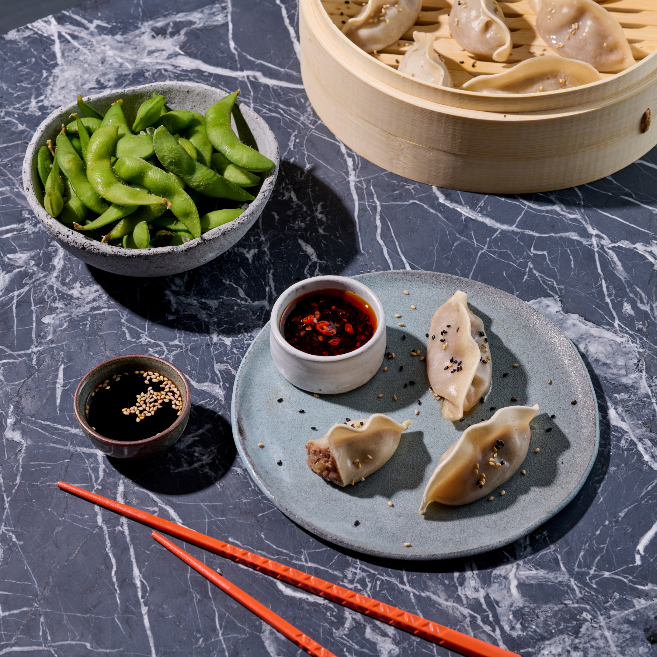 Dampfnudeln und Edamame auf Marmortisch, serviert mit Sojasauce und Chili-Dip, rote Essstäbchen daneben.