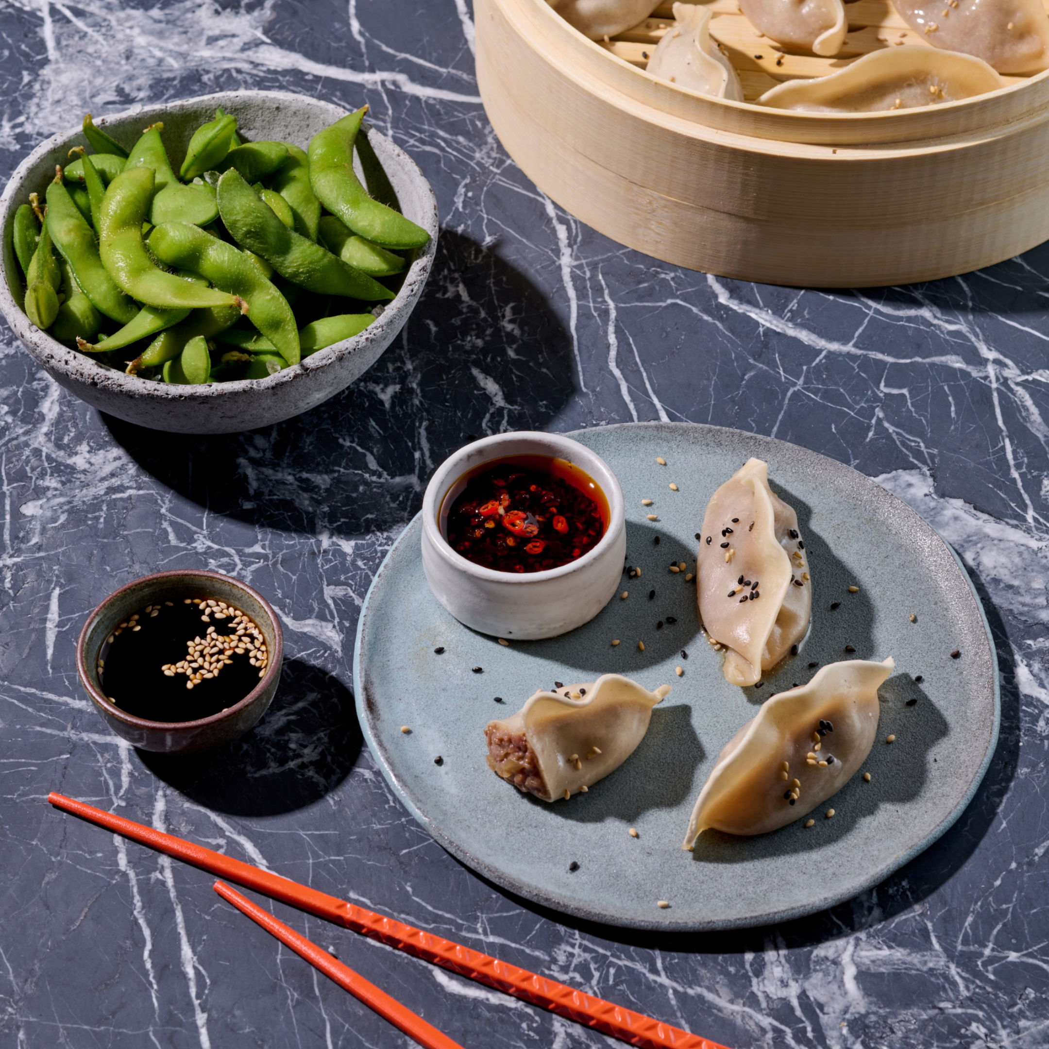 Dampfnudeln und Edamame auf Marmortisch, serviert mit Sojasauce und Chili-Dip, rote Essstäbchen daneben.