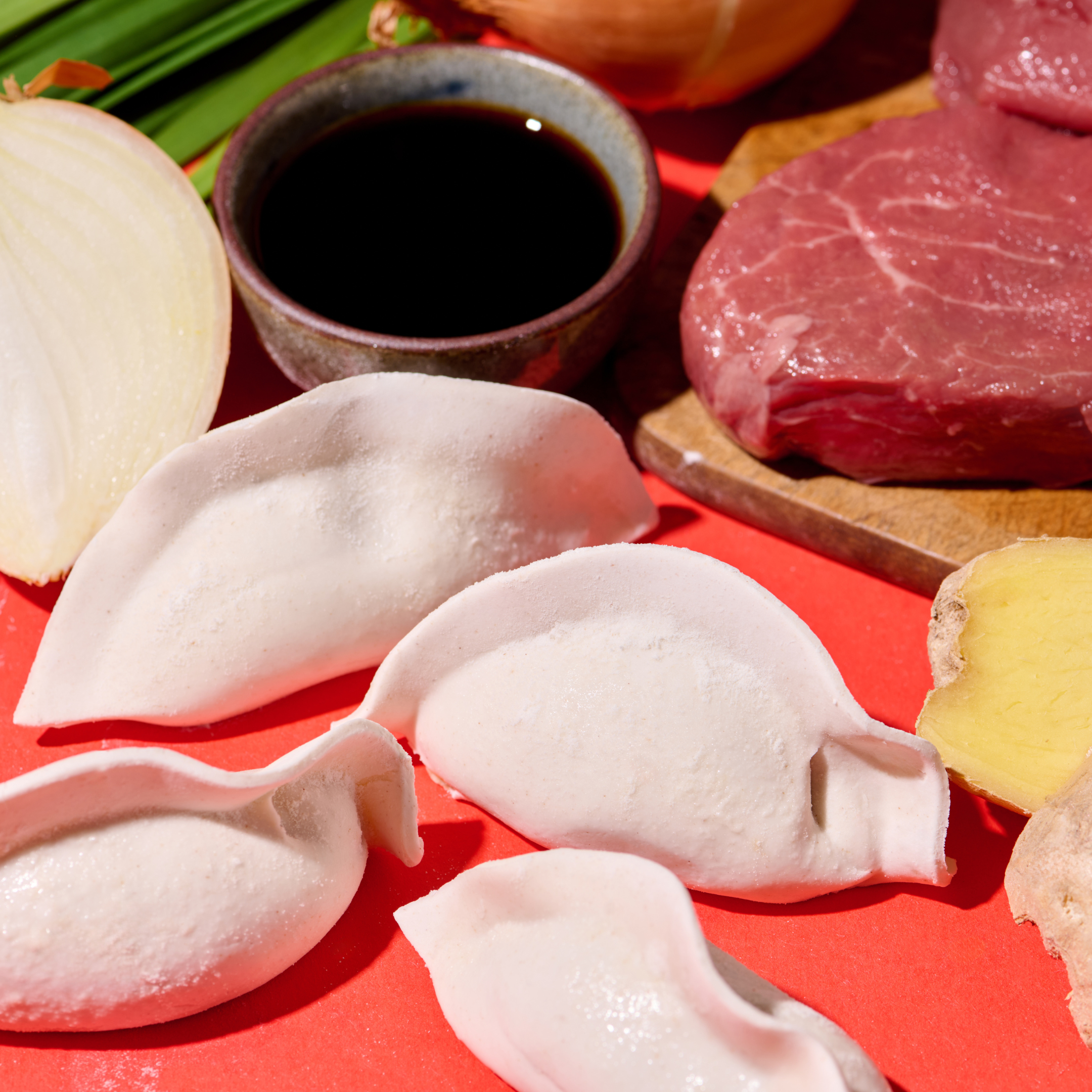 Rohes Fleisch, Zwiebel, Sojasauce, Teigtaschen und Ingwer auf einem roten Hintergrund arrangiert.