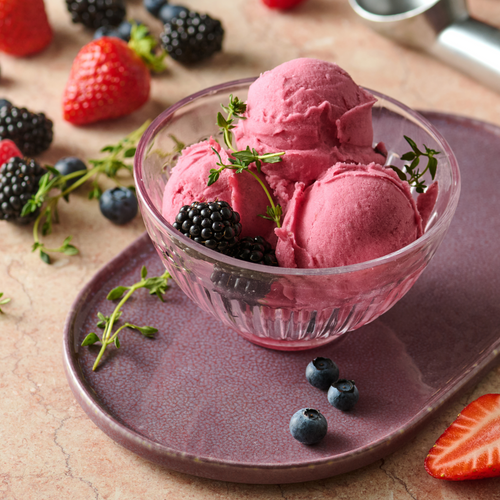 Drei Kugeln rosa Sorbet in einer Glasschüssel, garniert mit Beeren und Thymian, auf einem violetten Teller.