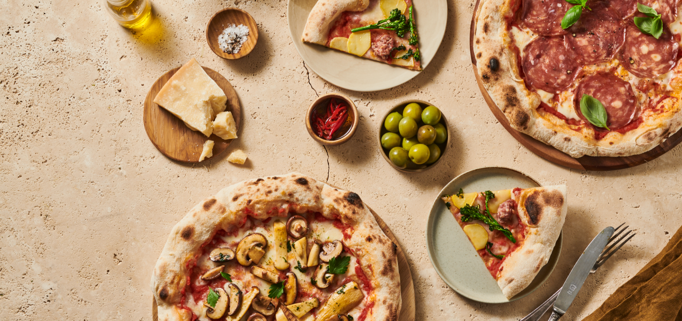 Verschiedene Pizzen mit Belägen wie Pilzen, Salami und Gemüse, serviert mit Parmesan, Oliven, Öl und Gewürzen.