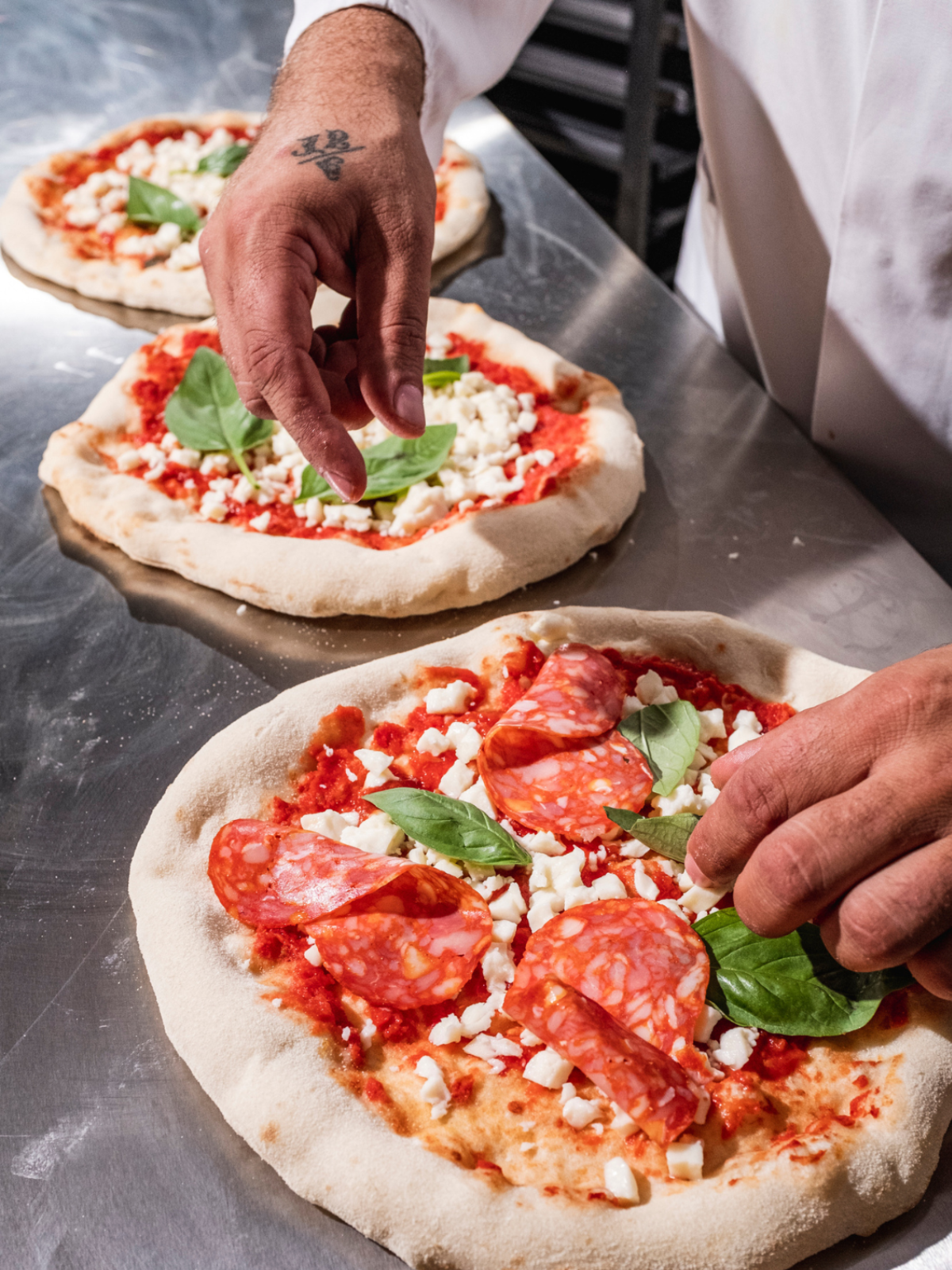 Ein Koch belegt Pizzen mit Salami, Käse und Basilikum auf einer Metalloberfläche.