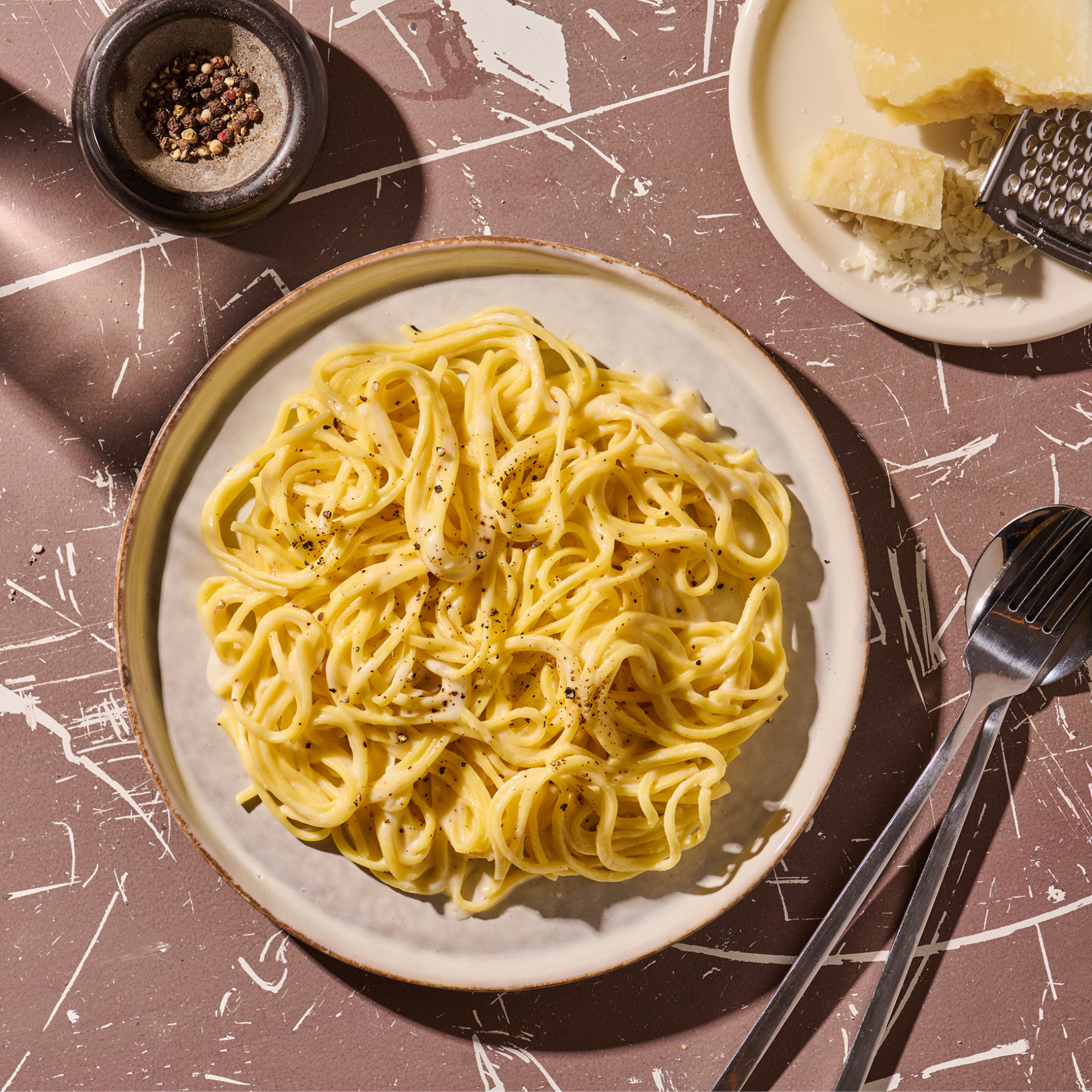 Ein Teller Spaghetti Carbonara mit schwarzem Pfeffer, daneben Pfefferkörner, Käse und Besteck auf einem Tisch.