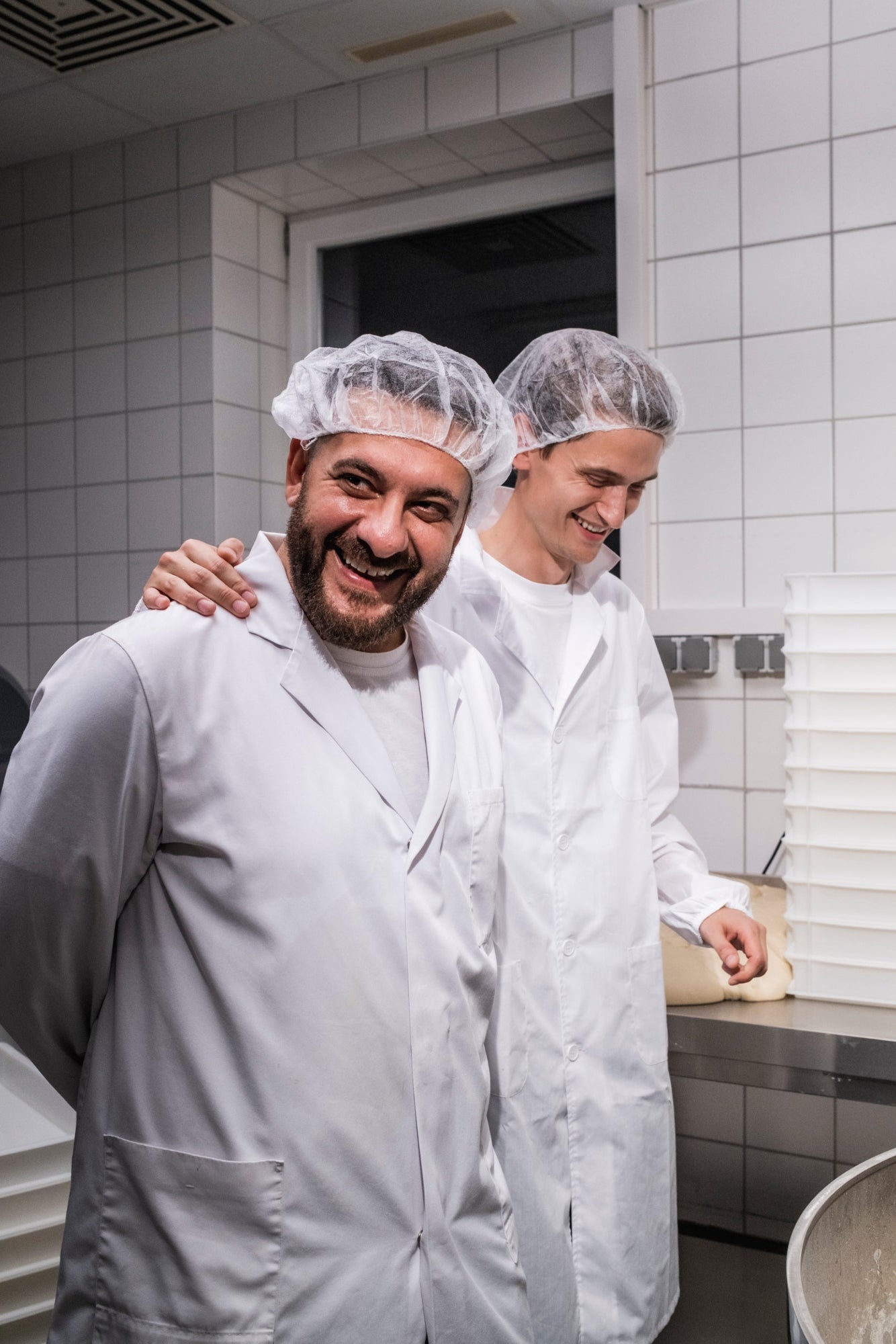 Zwei Männer in weißen Kitteln und Haarnetzen lächeln in einem Labor oder einer Bäckerei mit weißen Fliesenwänden.
