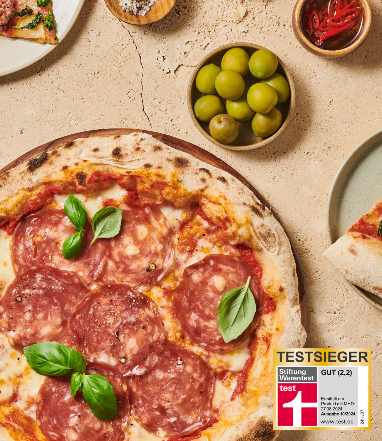 Pizza mit Salami und Basilikum auf hellem Untergrund, neben grünen Oliven und einem Testsieger-Label von Stiftung Warentest.