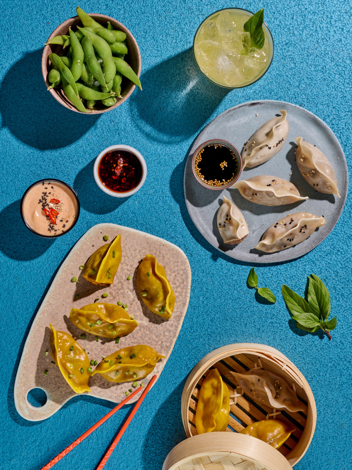 Verschiedene asiatische Gerichte wie Gyoza, Edamame, Saucen und ein Getränk auf einem blauen Hintergrund arrangiert.