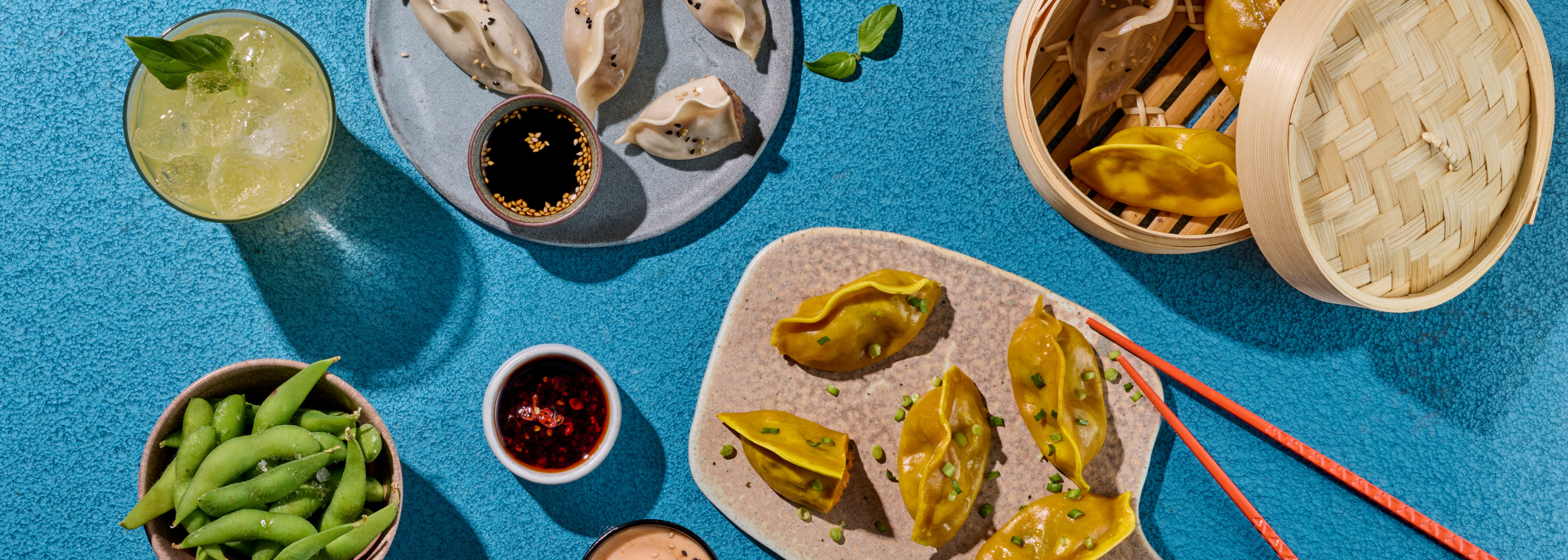 Asiatische Dumplings und Edamame mit Dippsaucen, einem Getränk und Essstäbchen auf einem blauen Tisch.
