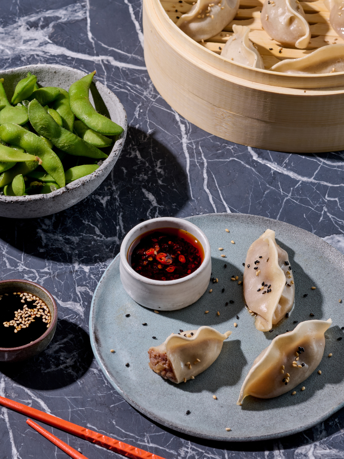 Ein Teller mit Dumplings und einer Schale Soße, umgeben von Edamame und Sojasoße auf einem Marmortisch.