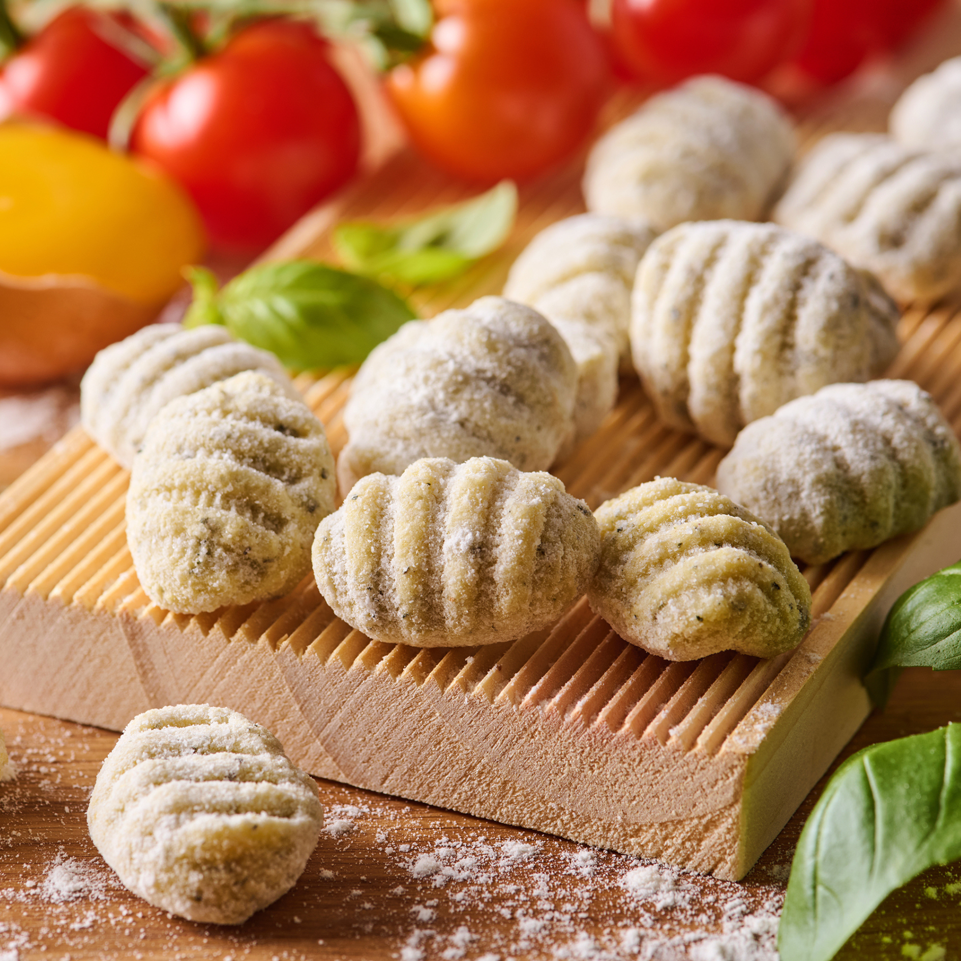 Gnocchi mit Mehl auf einem Holzbrett, umgeben von Basilikumblättern und bunten Tomaten im Hintergrund.